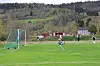 Fotballspillere på en grønn bane med mål til venstre og skogkledd bakgrunn.