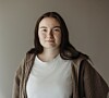 Portrett av en kvinne i hvit topp og brun cardigan foran en nøytral vegg.