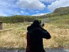 Person i mørke klær sikter med hagle mot et åpent landskap med skog og fjell.