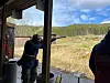 Personer skyter fra en overbygd standplass mot leiirduer i et åpent landskap.