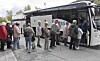 Folk står i kø ved en hvit buss med logo på en parkeringsplass.