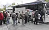 Folk står i kø ved en hvit buss med logo på en parkeringsplass.