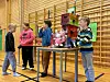 Barn står rundt et bord med en selvkonstruert robot med bamser i en gymsal med ribbevegger.