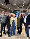 Fem personer står side om side innendørs i et lyst lokale med skjerm og skilting bak dem.