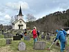 Personer arbeider med rydding på en kirkegård med gravsteiner, liten kirke og skog i bakgrunnen.