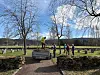 Personer rydder og arbeider på en gravlund med gravsteiner, trær og en tilhenger i solskinn.