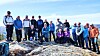 En gruppe personer står og sitter på en steinete fjelltopp i solskinn.