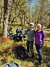 To kvinner står foran en gruppe turgåere med staver og sekker i en skog på en solrik dag.
