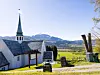 Hvit kirke med spisst tårn i et landlig landskap med fjell og grønne jorder.