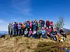 Stor gruppe mennesker står samlet på et gresskledd fjell med hav og fjell i bakgrunnen.
