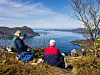Tre personer sitter på en gresskledd høyde med utsikt over fjord og fjell under klar himmel.