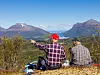 To personer sitter på en ås og peker utover et landskap med fjell, skog og åpen himmel.