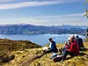 To personer sitter på en gresskledd ås med utsikt mot innsjø og fjell under klar himmel.