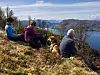 Tre personer og en hund sitter på en høyde med utsikt over fjord, fjell og trær i høstfarger.
