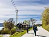 To turgåere på en vei ved vannet med hus, trær og kraftlinjer langs traseen.