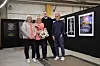 Fire personer står foran innrammede kunstverk i et galleri med svarte vegger.