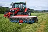 Rød traktor med slåmaskin kjører over en grasmark i et kupert jordbruksområde.