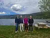 Fire personer står ved benker langs en tursti ved vannet, med fjell og overskyet himmel bak.