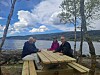 Fire personer sitter ved et piknikbord ved vannet med fjell og skyer i bakgrunnen.