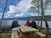 Fire personer sitter ved et piknikbord ved vannet med fjell og skyer i bakgrunnen.