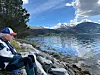 En mann sitter på steiner ved vannkanten med innsjø og fjell i bakgrunnen.