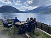 Fire personer sitter rundt et bord på en terrasse ved en innsjø med fjell i bakgrunnen.