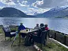 Fire personer sitter rundt et bord på en terrasse ved en innsjø med fjell i bakgrunnen.