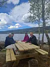 Fire personer sitter ved et trebord ved en innsjø med fjell og trær i bakgrunnen.