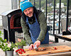 En mann i lue og forklær skjærer pizza ved et bord ved en vedfyrt ovn.