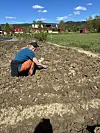 Person på huk i en jordhaug på et gårdsbruk med hus og åkre i bakgrunnen.