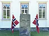 Steinmonument foran hvit kirkebygning med to norske flagg på hver side.