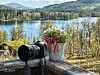 Kamera og blomsterkrukke med bånd i fargene til det norske flagget, på en benk foran en innsjø og skogkledde fjell.