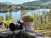 Kamera og blomsterkrukke med bånd i fargene til det norske flagget, på en benk foran en innsjø og skogkledde fjell.