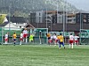 Ungdommer spiller fotball foran et mål på en kunstgressbane med fjell og bygninger bak.