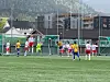 Ungdommer spiller fotball foran et mål på en kunstgressbane med fjell og bygninger bak.