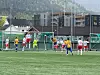 Ungdommer spiller fotball foran et mål på en grønn kunstgressbane med fjell og bygninger i bakgrunnen.