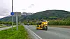 Gul motorsykkel kjører på en vei med grønne åser og snødekte fjell i bakgrunnen.