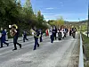 Et korps og flere folk går i et 17. maitog langs en vei omkranset av grønt landskap.