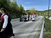 Et lite 17. mai-tog med musikanter, barn og voksne går langs en vei i solskinn.