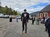 En mann med krykker står på en grusplass foran et rødt hus, med flere personer i bakgrunnen.