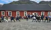 Flere personer trekker i et tau på en grusplass foran en rød bygning med fjell bak.