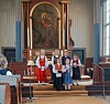 Et lite barnekor står samlet foran alteret i en kirke med et stort religiøst maleri bak dem.