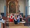 Et lite barnekor står samlet foran alteret i en kirke med et stort religiøst maleri bak dem.