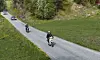 En person kjører moped foran to biler på en svingete vei gjennom grønt landskap.