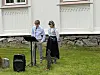 To personer står på gresset ved et mikrofonstativ foran en lys kirkevegg