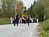 Et 17. mai-tog med barn og voksne går langs en vei omgitt av trær og vårblomster.