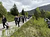 Et barnetog i festklær går langs en vei med grønne busker og fjell i bakgrunnen.