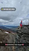 Norsk flagg står på en steinrygg med vid utsikt over fjell og dal under en skyet himmel.