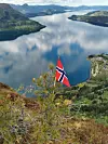 Norsk flagg i forgrunnen med fjord, fjell og skog i bakgrunnen.