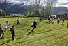 Mennesker beveger seg over en gresslette med fjell og vann i bakgrunnen.